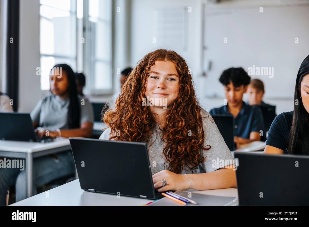Portrait d'étudiant adolescent aux cheveux bouclés souriants assis avec un ordinateur portable près du bureau dans la salle de classe à l'école Banque D'Images
