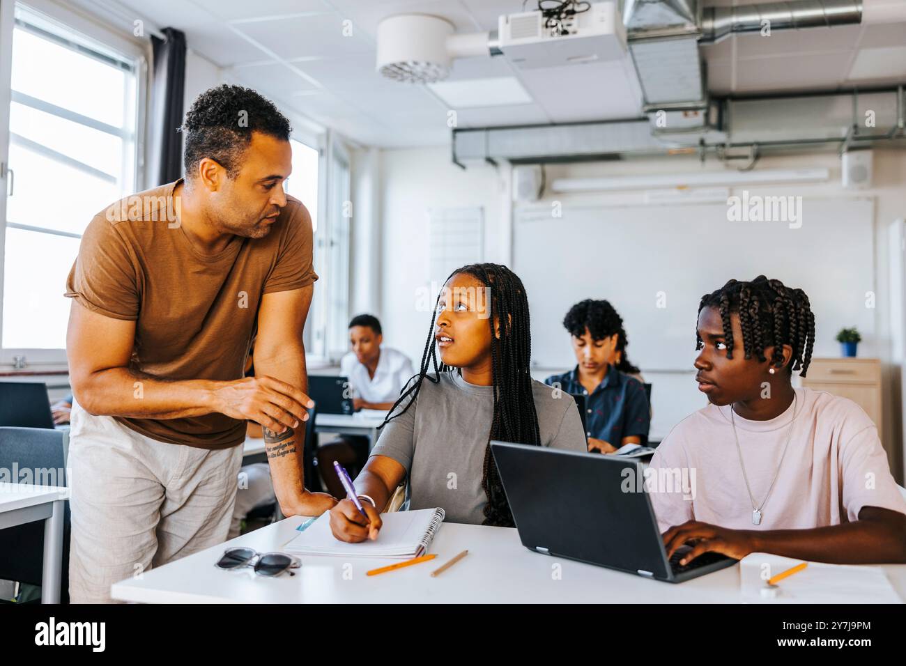 Professeur masculin aidant des étudiants adolescents assis avec un ordinateur portable dans la salle de classe au collège Banque D'Images