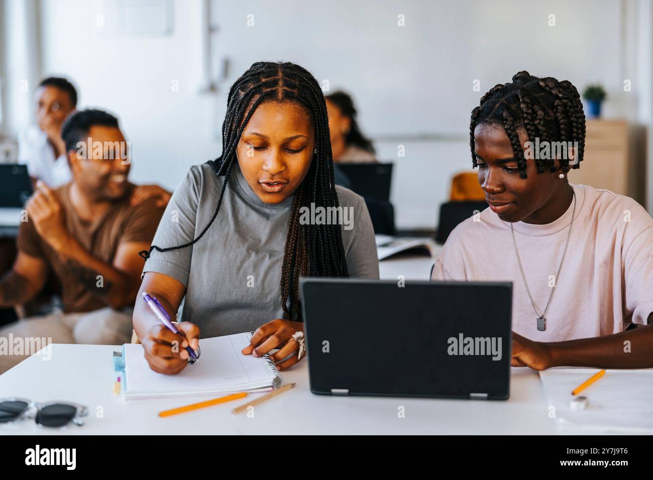 Fille de cheveux tressés écrivant tout en étant assise à côté d'un camarade de classe masculin près du bureau dans la salle de classe à l'école Banque D'Images