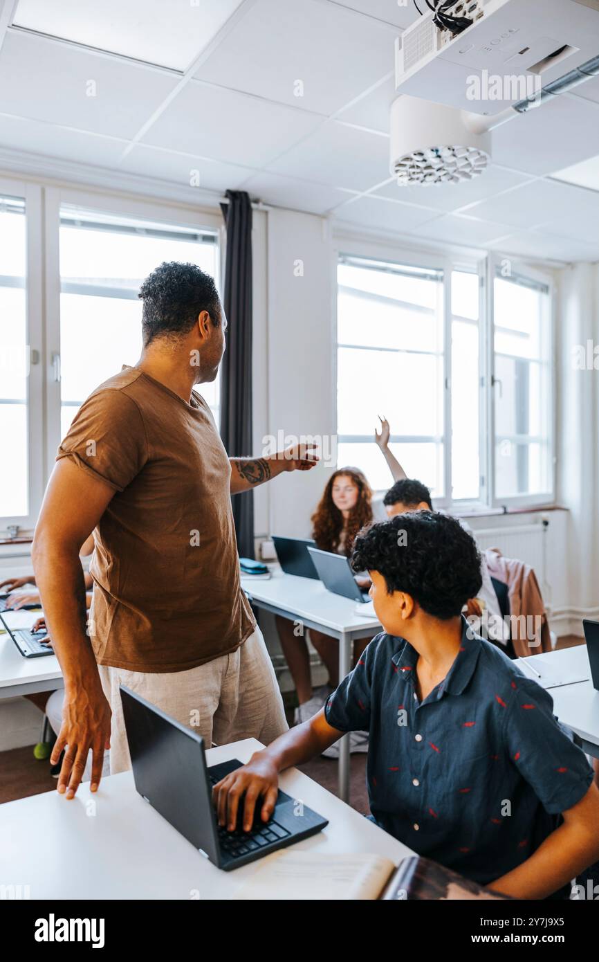 Professeur masculin aidant les étudiants à étudier à travers un ordinateur portable tout en étant assis à un bureau dans la salle de classe Banque D'Images