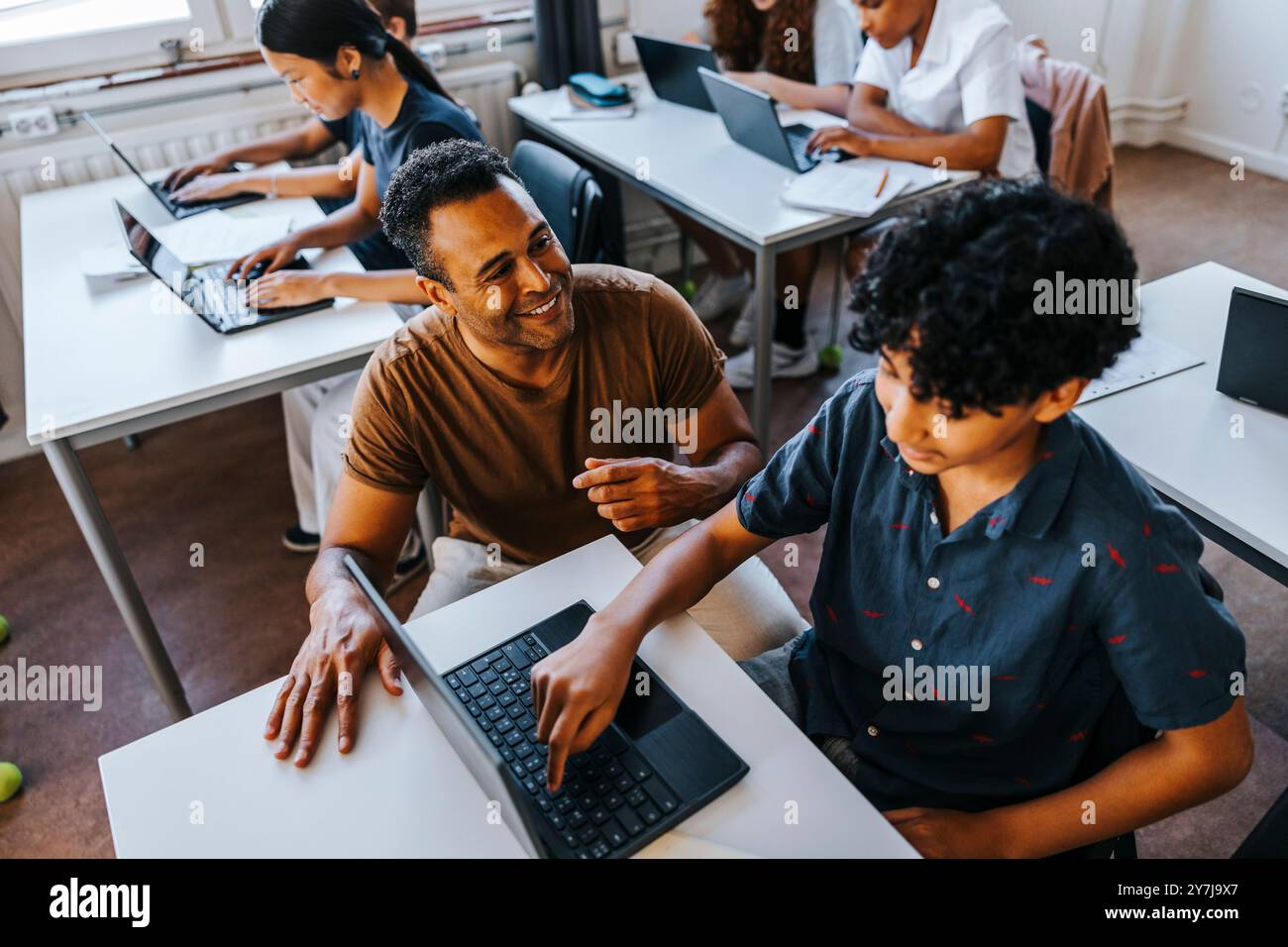 Vue en grand angle d'un professeur heureux accroupi près d'un étudiant du secondaire utilisant un ordinateur portable tout en étant assis dans la salle de classe à l'école Banque D'Images