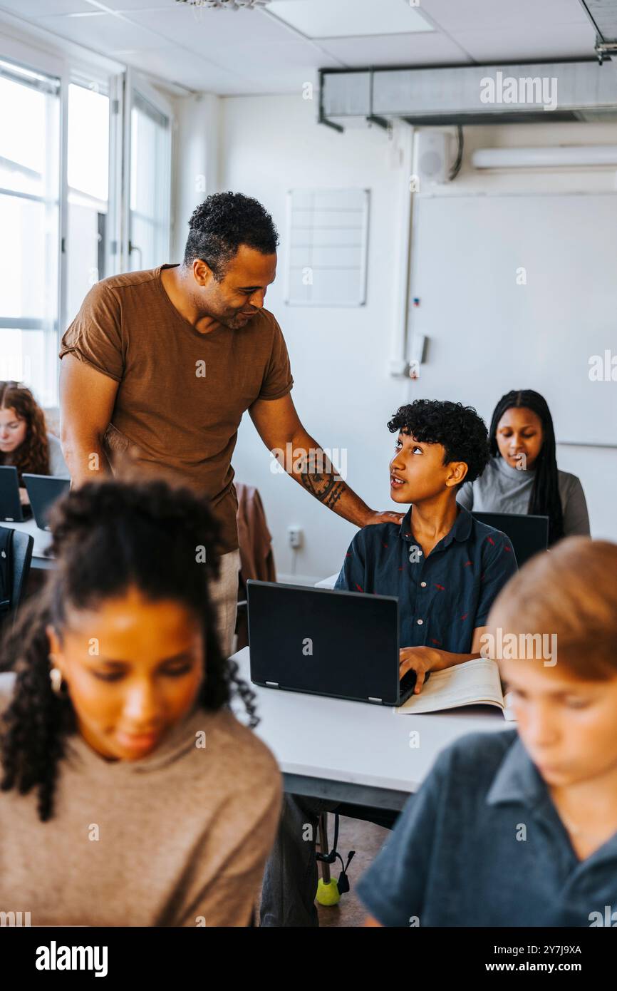 Professeur de sexe masculin guidant l'étudiant adolescent se trouvant avec un ordinateur portable dans une salle de classe au collège Banque D'Images