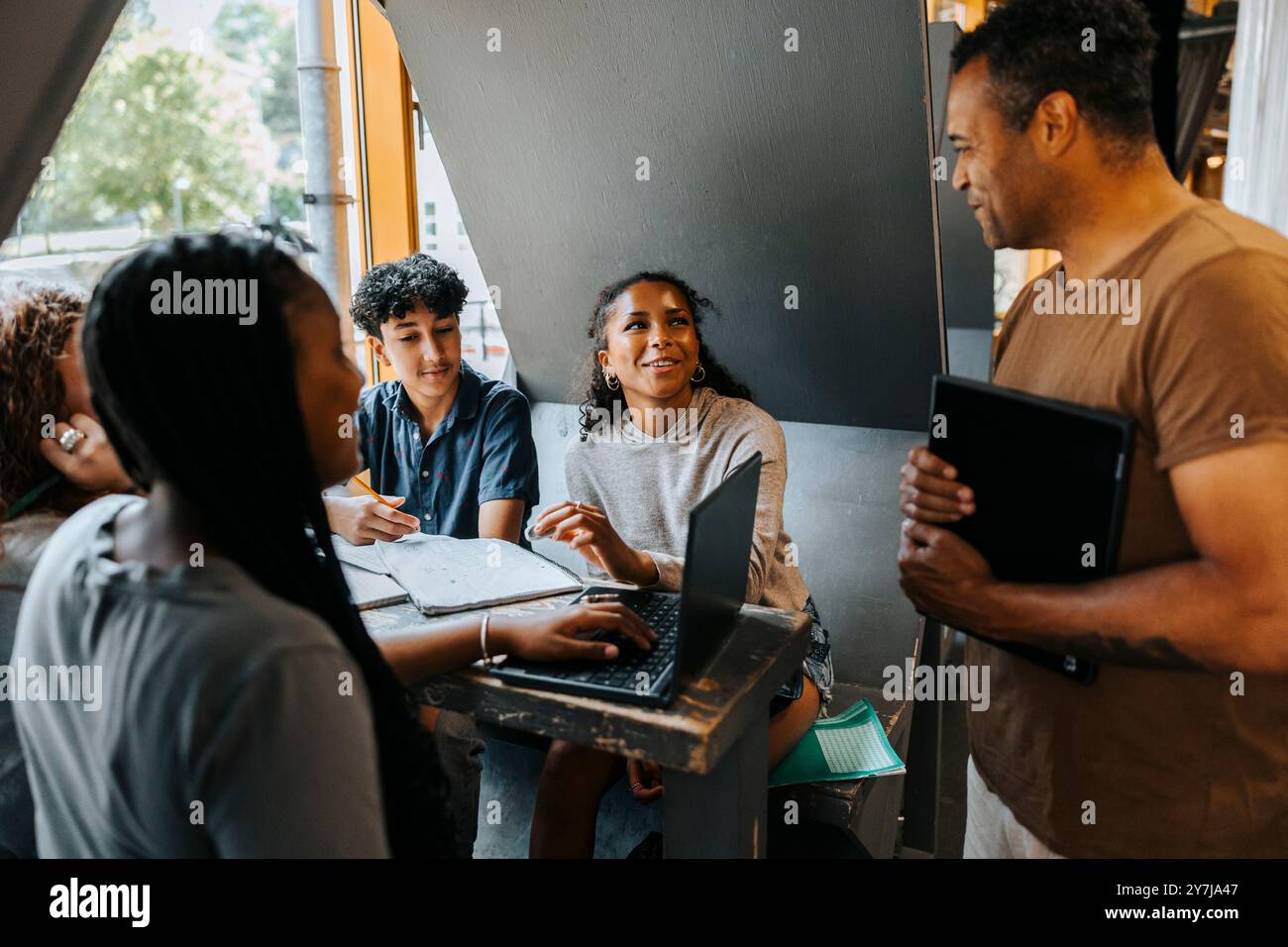 Heureux professeur aidant le groupe d'étudiants étudiant par ordinateur portable tout en étant assis à la cafétéria au collège Banque D'Images