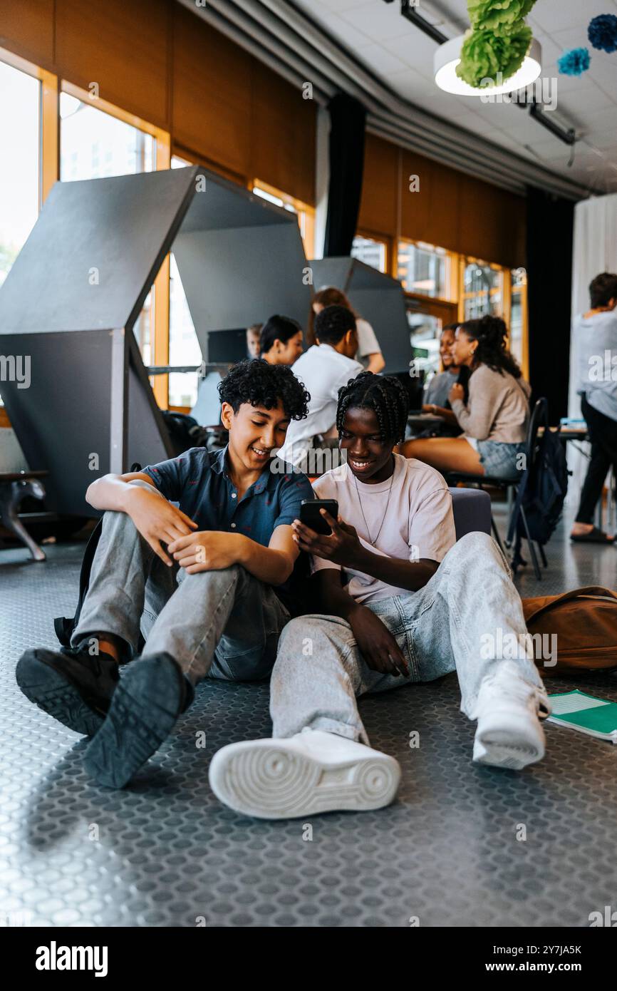 Heureux amis masculins utilisant un téléphone intelligent tout en étant assis dans la cafétéria à l'école Banque D'Images