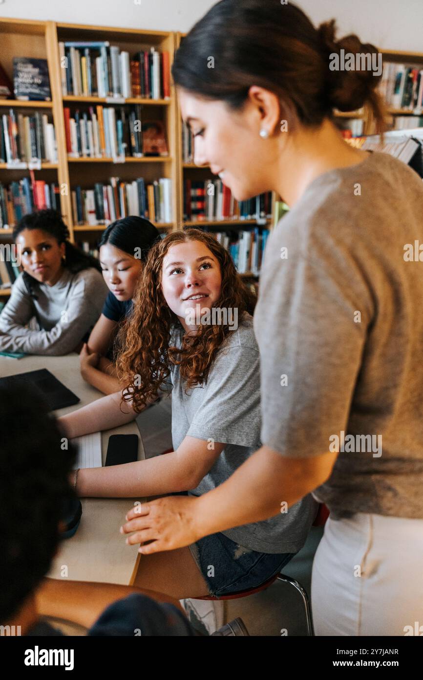 Bibliothécaire féminine aidant un élève adolescent assis près d'une table dans la bibliothèque à l'école Banque D'Images