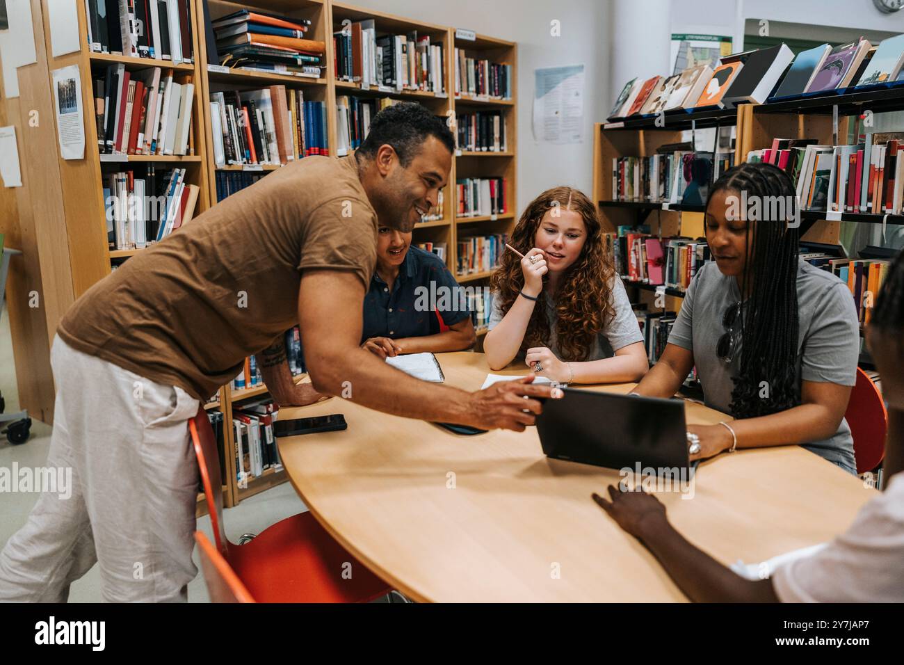 Heureux bibliothécaire masculin aidant les élèves adolescents assis près de la table dans la bibliothèque à l'école Banque D'Images