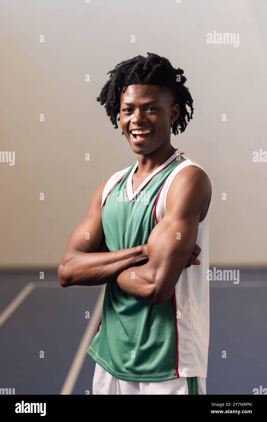 Joueur de basket-ball souriant en maillot vert debout avec les bras croisés dans la salle de gym Banque D'Images