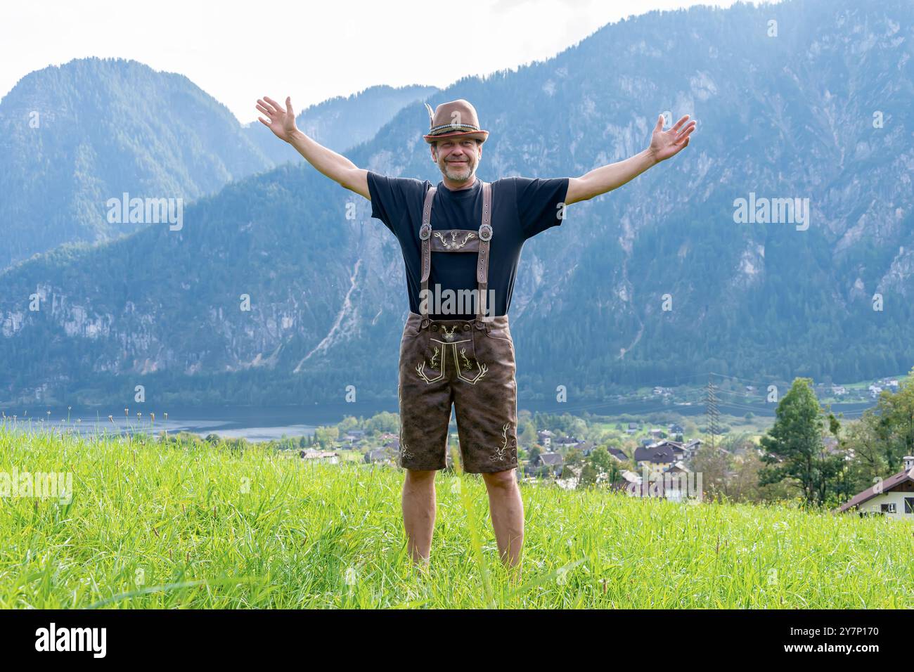 Un agriculteur autrichien en tenue nationale se dresse sur un fond de montagnes et une petite ville, les bras étendus. Banque D'Images