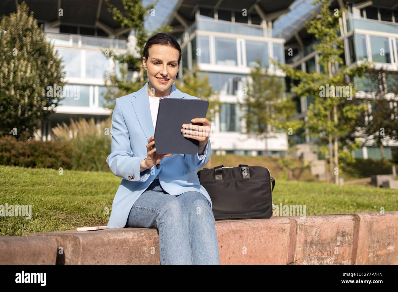 Heureuse femme d'affaires utilisant une tablette numérique à l'extérieur profitant d'une journée ensoleillée avec des immeubles de bureaux modernes en arrière-plan Banque D'Images