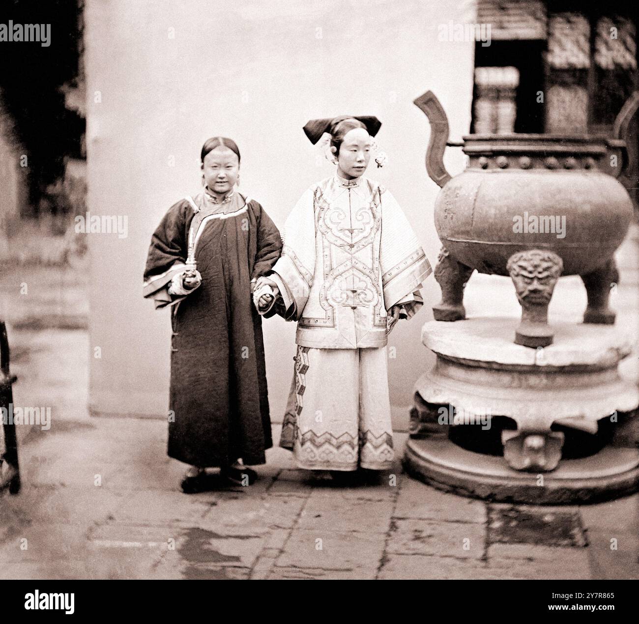 Photo vintage d'une femme mandchou avec sa femme de ménage debout à côté d'un brûleur en bronze, Pékin. Zhili du Nord (Pei-chih-Li ou province de Pékin), Chine. Par John Thomson, 1869. Deux femmes debout à côté d'un brûleur d'encens, l'une tenant le bras de l'autre. La dame porte un yunjian (col nuageux). Alors que la femme de chambre regarde directement la caméra, sa maîtresse semble sinistre, suggérant que Thomson essayait de s'éloigner de la tradition formelle du portrait, essayant de dépeindre ses sujets comme des individus. Cela montre aussi qu'il essayait de transmettre le rôle ambigu d'une servante mandchou : elle ressemblait plus à une compagne Banque D'Images