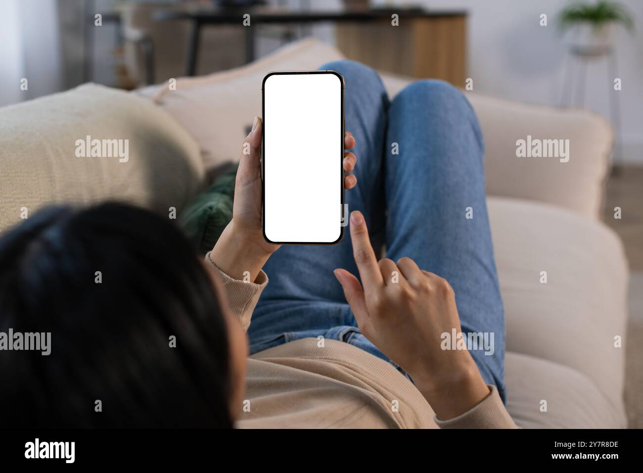 Une jeune femme asiatique méconnaissable tenant une maquette de smartphone avec un écran blanc vierge couché sur le canapé confortable à la maison, point de vue Banque D'Images