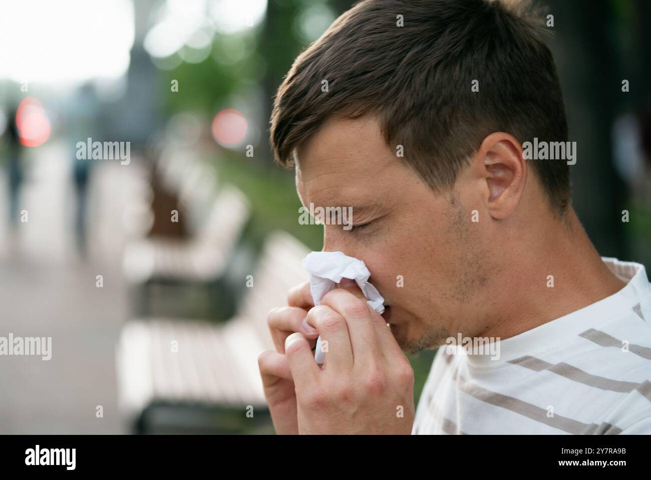 Gros plan d'un homme qui souffle son nez dans un mouchoir alors qu'il était assis dans un parc, mettant en évidence l'hygiène et les allergies saisonnières. Banque D'Images