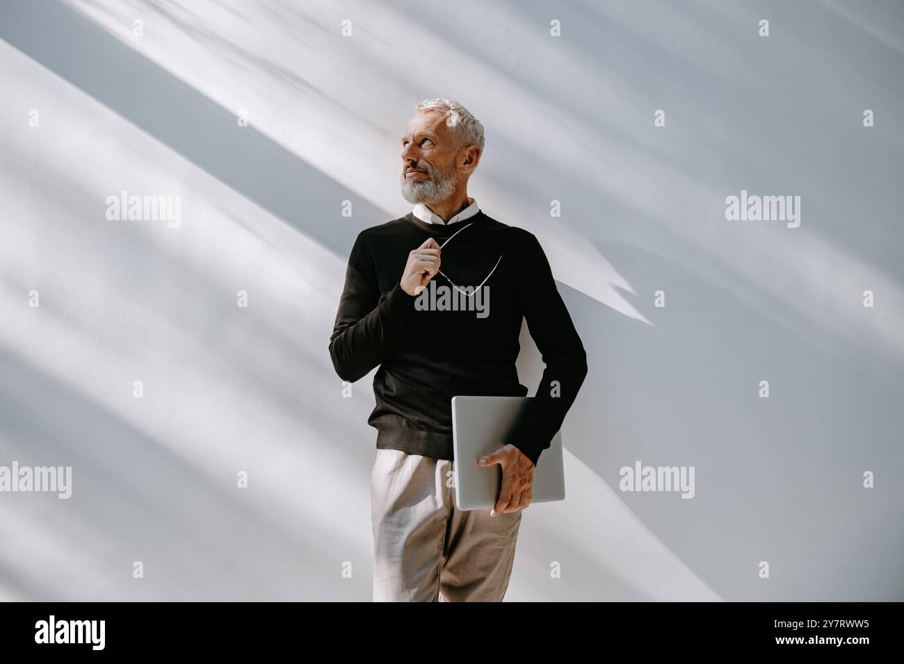 Portrait de l'homme de cheveux gris confiant dans une tenue décontractée intelligente tenant un ordinateur portable tout en se tenant debout contre un mur Banque D'Images