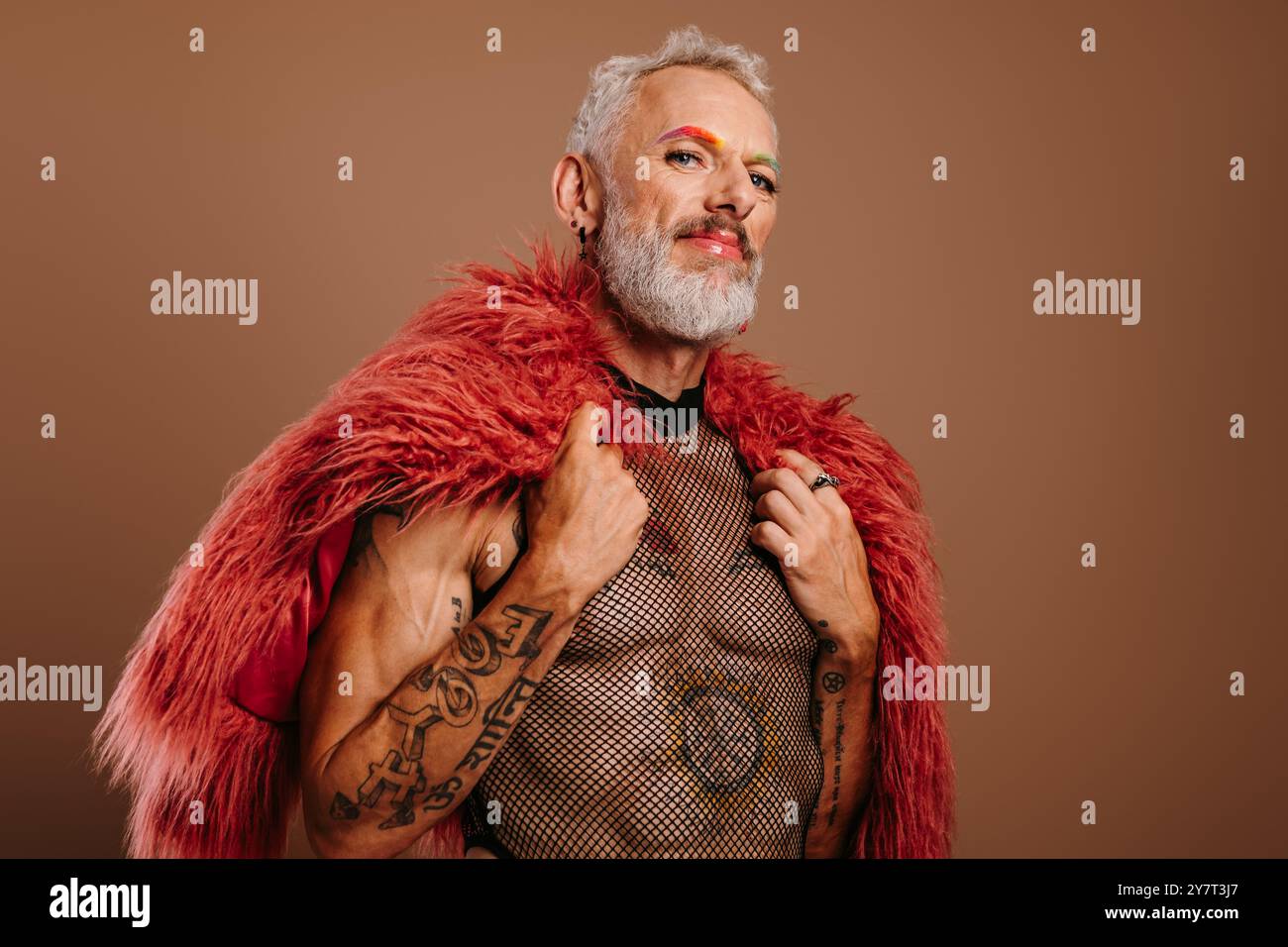 Homme gay mature avec des sourcils colorés arc-en-ciel portant un manteau moelleux sur fond brun Banque D'Images