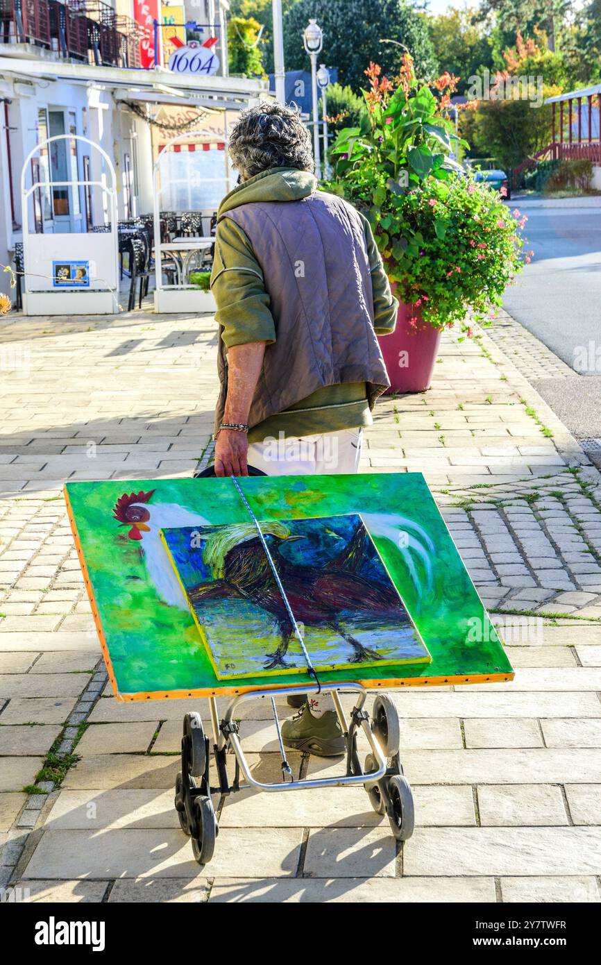 Femme artiste amateur tirant chariot avec des œuvres d'art en toile le long du trottoir - la Roche Posay, Vienne (86), France. Banque D'Images