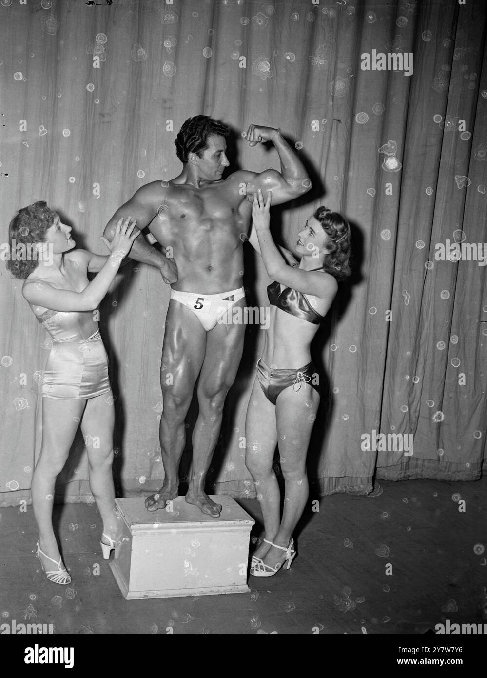 Les muscles puissants du Mr Universe de 1952 viennent pour l'admiration d'une jolie paire, Joyce Martin (à gauche), de Plymouth, Devon, et Thelma Nowlan, 17 ans, également de Plymouth, après le jugement pour le titre lors d'un concours au Scala Theatre. M. Universe est un Espagnol, Juan Ferrero, qui vit à Bordeaux, France.12 juillet 1952 Banque D'Images