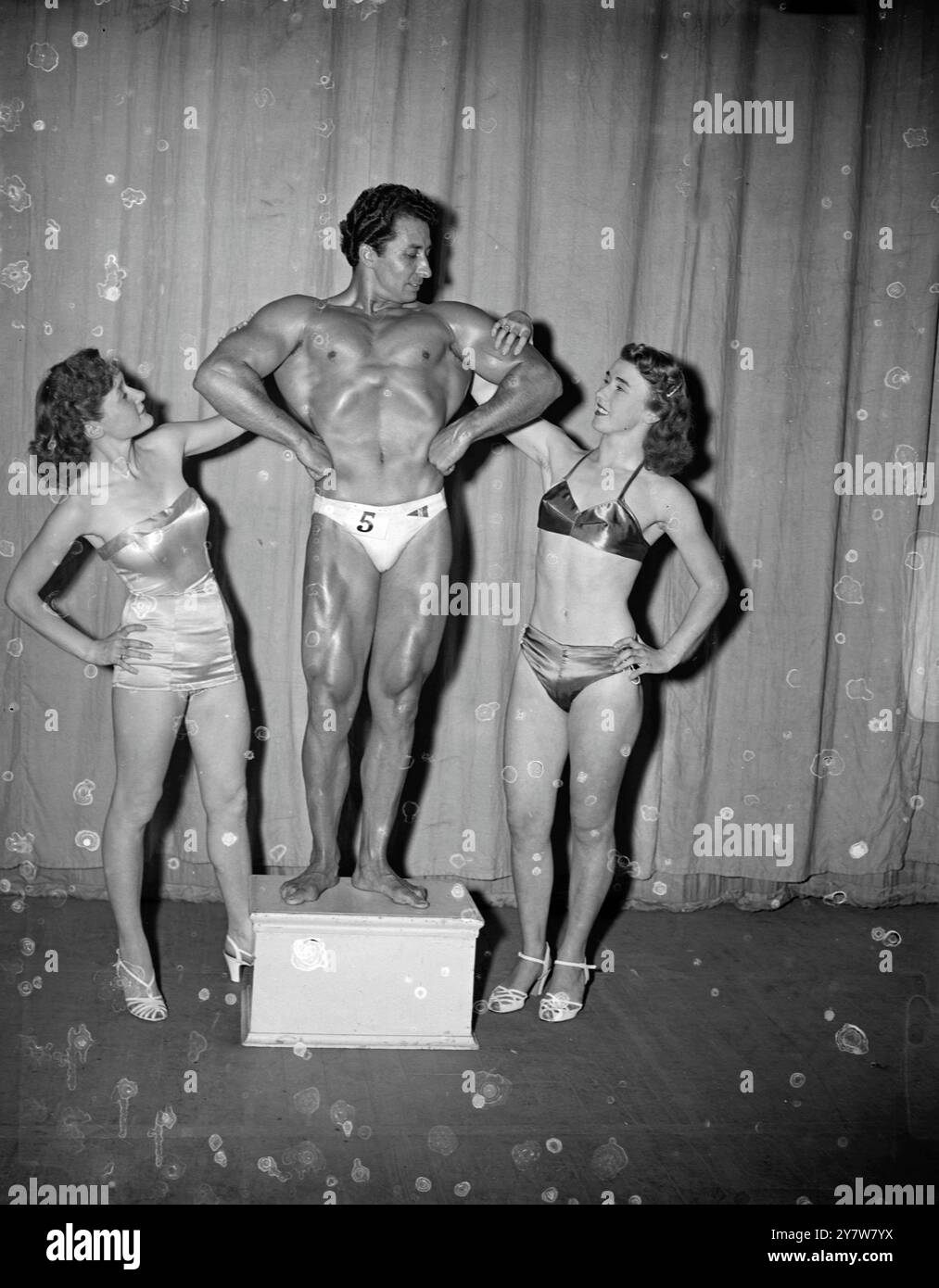 Les muscles puissants du Mr Universe de 1952 viennent pour l'admiration d'une jolie paire, Joyce Martin (à gauche), de Plymouth, Devon, et Thelma Nowlan, 17 ans, également de Plymouth, après le jugement pour le titre lors d'un concours au Scala Theatre. M. Universe est un Espagnol, Juan Ferrero, qui vit à Bordeaux, France.12 juillet 1952 Banque D'Images