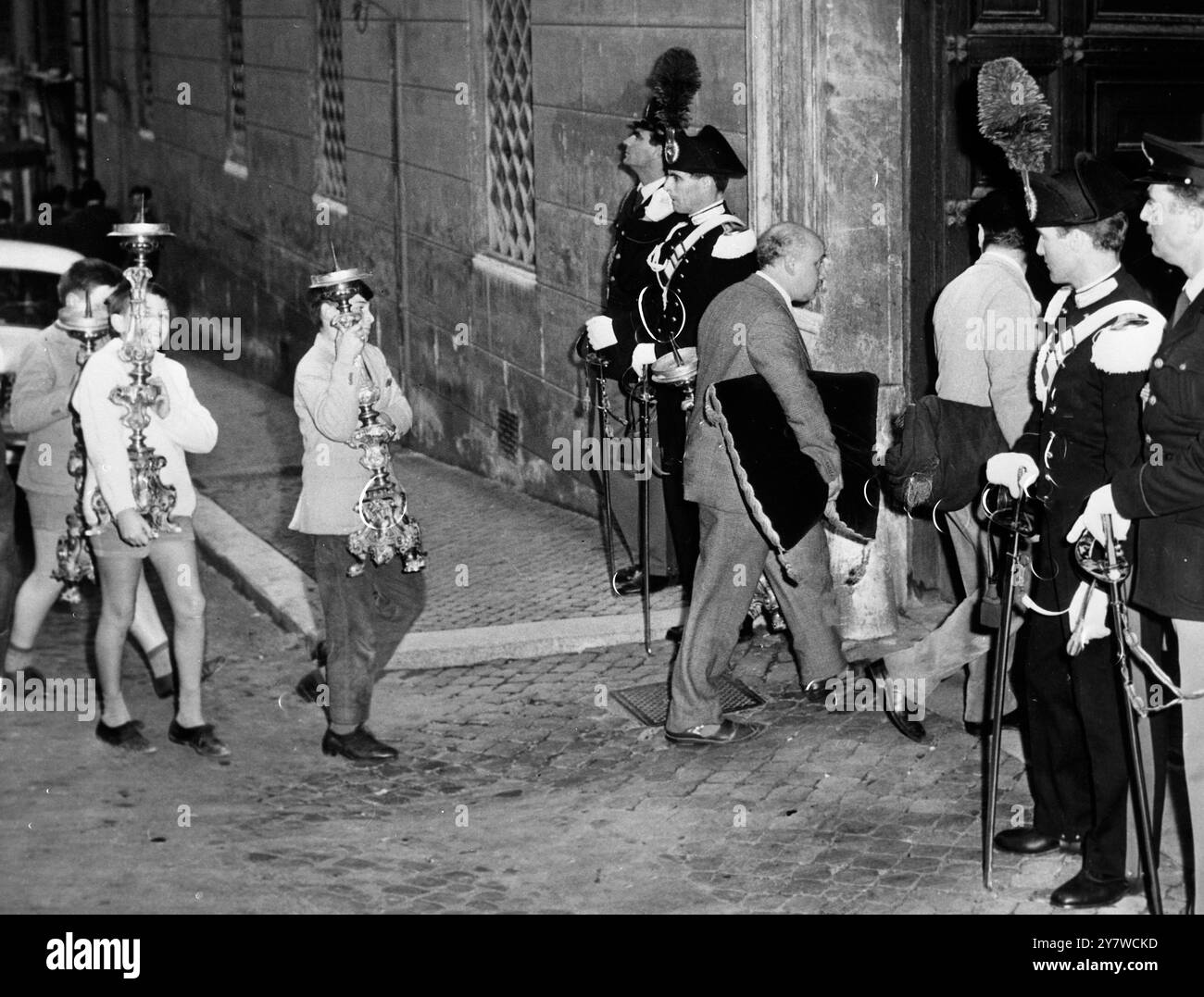 Des carabiniers en uniforme complet ( chapeaux à plumes ) et des policiers montent la garde à la porte du Palais Dataria à Rome hier , tandis que des draps noirs et des bougies sont amenés à l'intérieur , ce dernier devant être placé sur les côtés du corps du cardinal Federico Tedeschini , décédé le 2 novembre 1959 et se trouvant en état au palais . Le Cardinal est décédé à l'âge de 86 ans. Sa mort réduit le nombre de cardinaux à 71. - 4 novembre 1959 Banque D'Images