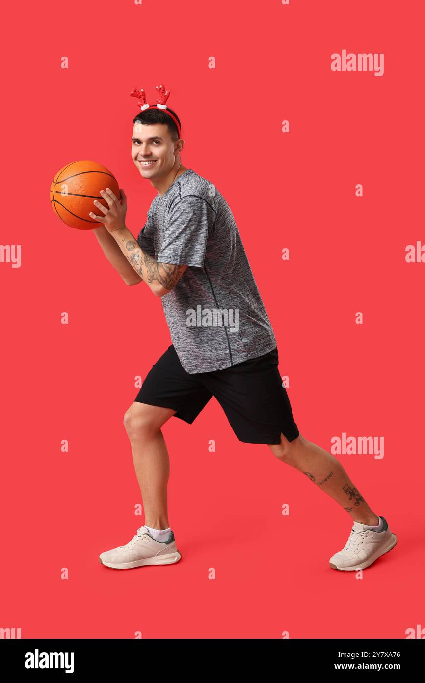 Jeune homme heureux dans des cornes de renne jouant au basket-ball sur fond rouge Banque D'Images