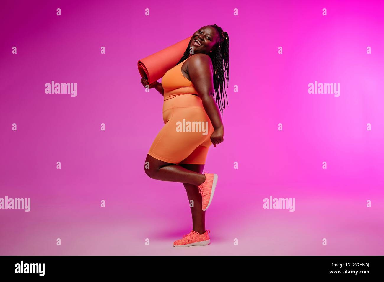 Jeune femme voluptueuse active dans des vêtements de sport portant un tapis d'exercice et souriant sur fond rose Banque D'Images