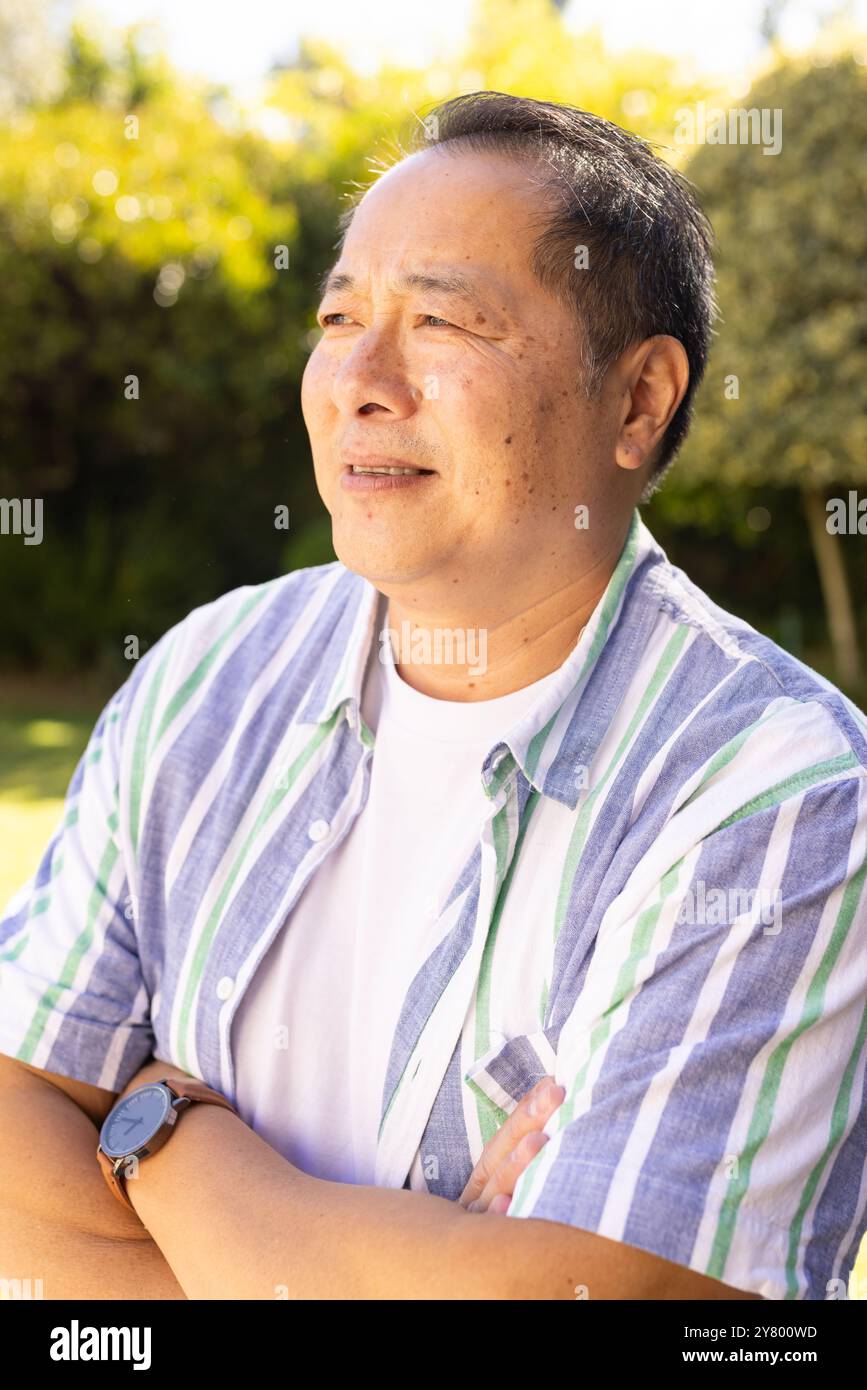 Homme d'âge moyen asiatique souriant debout à l'extérieur, croisant les bras et regardant loin, profitant d'une journée ensoleillée Banque D'Images