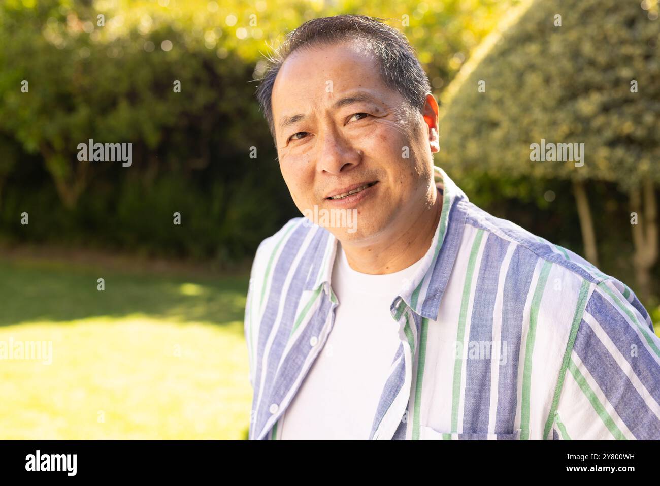 Souriant homme d'âge moyen asiatique en chemise rayée profitant d'une journée ensoleillée à l'extérieur dans le jardin Banque D'Images