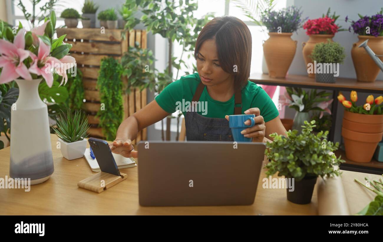 Une femme concentrée utilise un ordinateur portable et un téléphone portable dans un magasin de fleurs intérieur entouré de plantes vibrantes. Banque D'Images