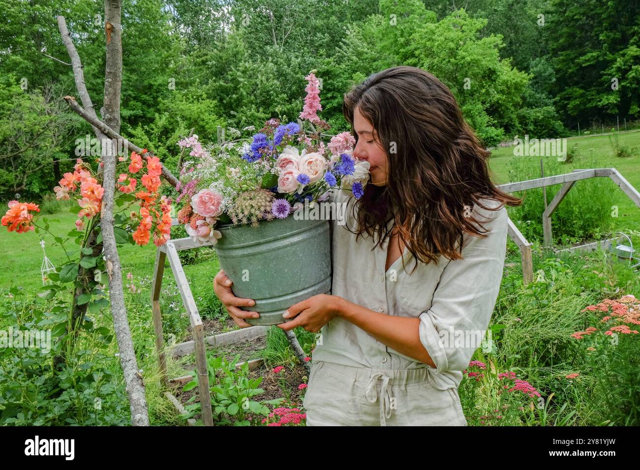 Une femme dans une robe légère sourit alors qu'elle tient un seau rempli de fleurs colorées dans un jardin luxuriant. Banque D'Images