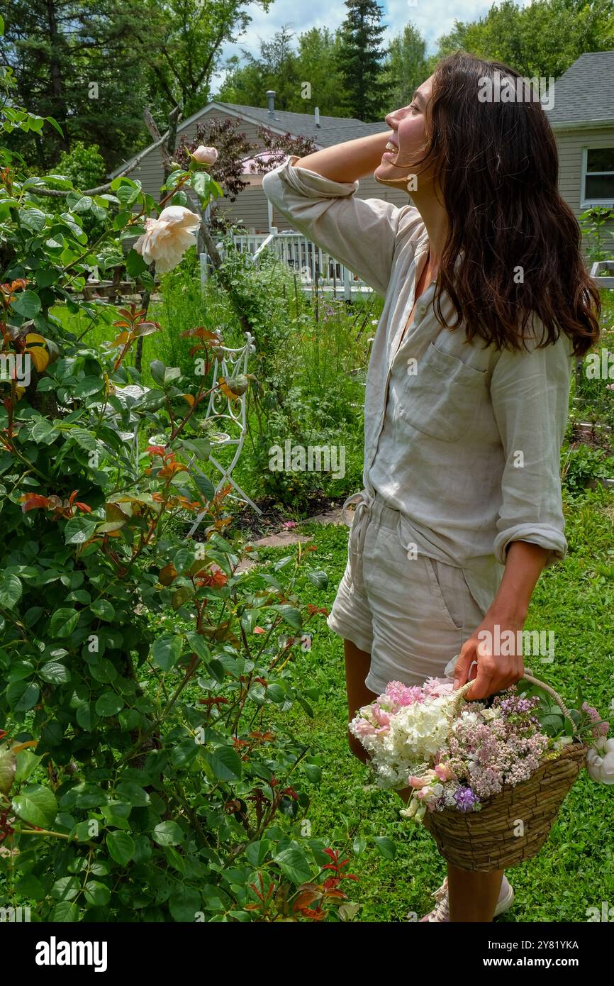 Femme dans une robe blanche profitant du parfum des fleurs dans un jardin luxuriant tenant un panier de fleurs fraîches. Banque D'Images