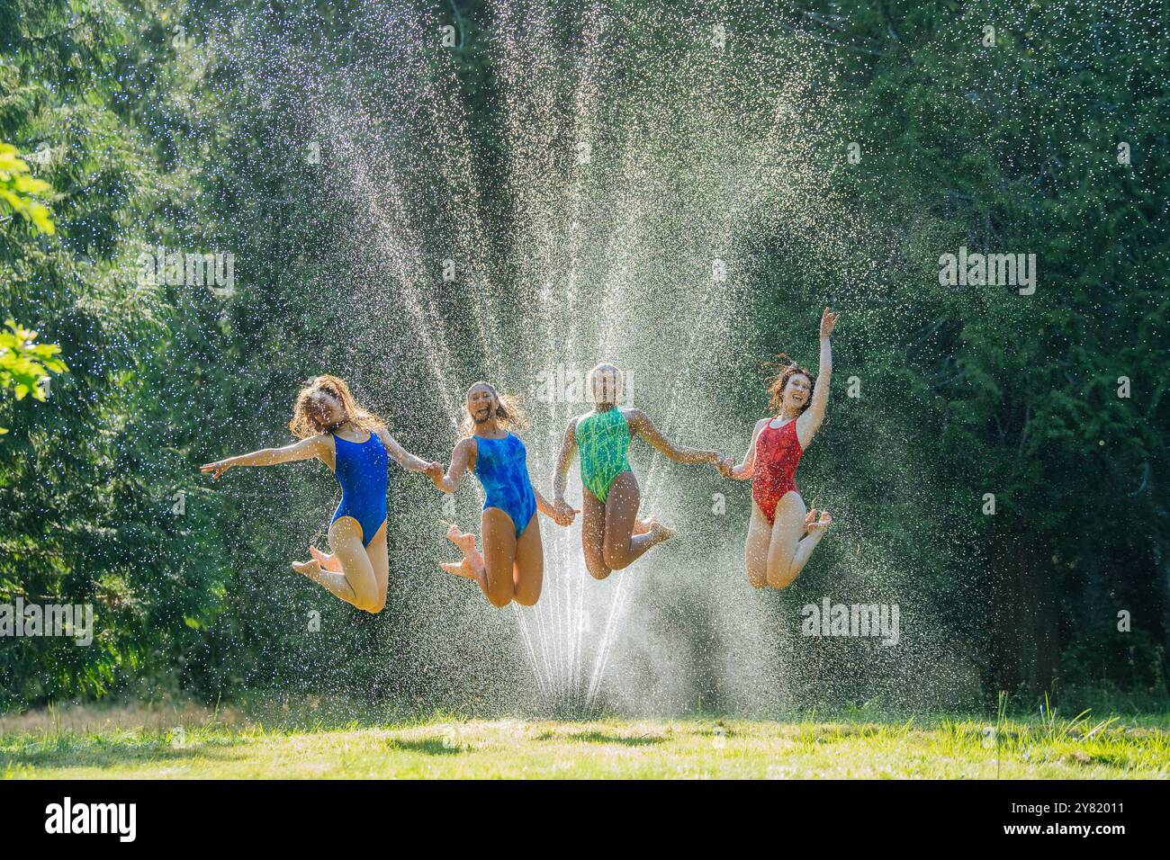 Joyeux groupe d'amis sautant et s'amusant sous un arroseur d'eau dans un parc par une journée ensoleillée. Banque D'Images