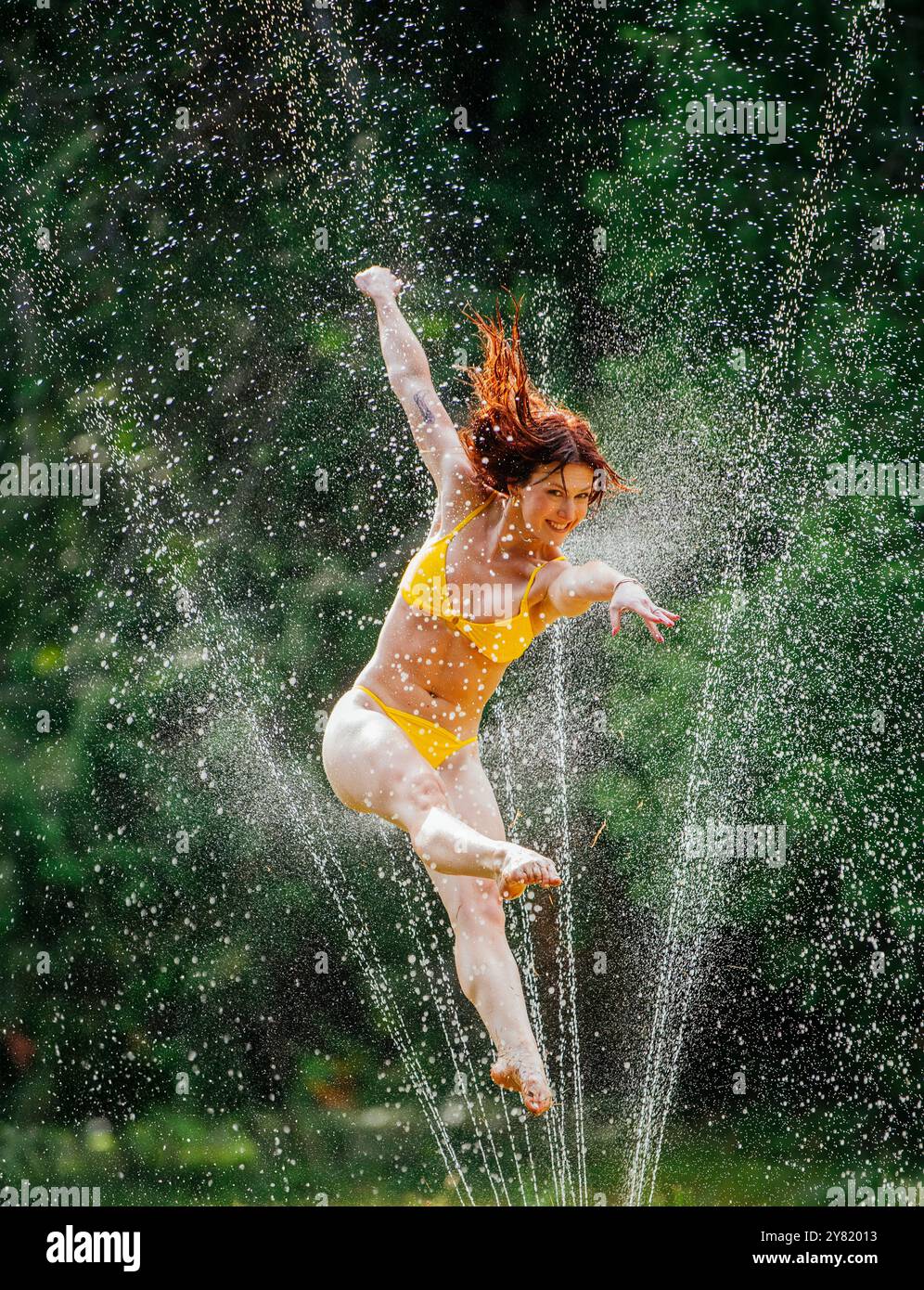 Femme joyeuse sautant par-dessus les gicleurs d'eau dans un jardin par une journée ensoleillée. Banque D'Images