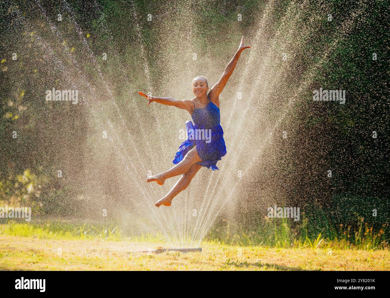 Femme joyeuse dans une robe bleue sautant à travers l'eau d'arrosage au soleil. Banque D'Images