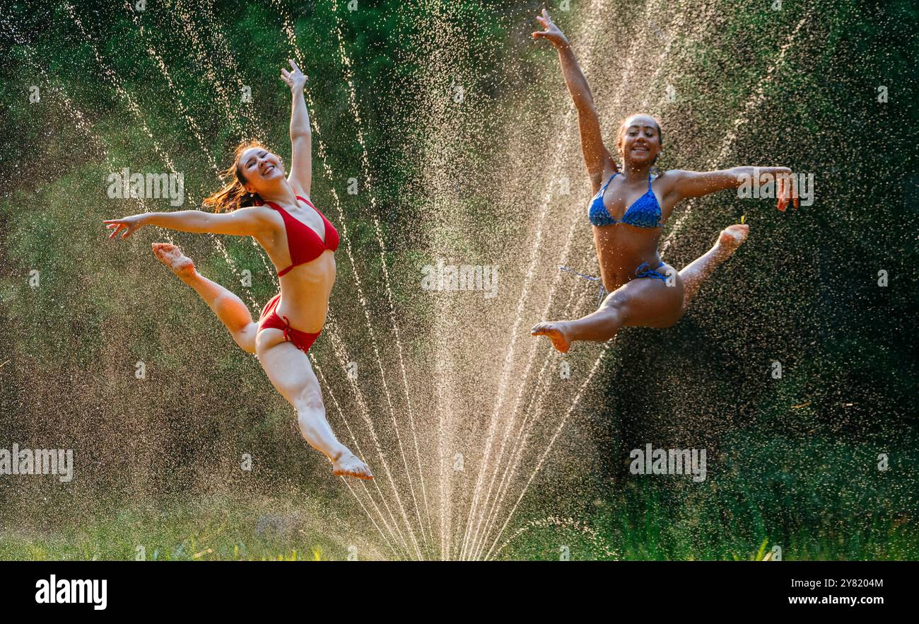 Deux femmes sautent joyeusement à travers des gicleurs d'eau par une journée ensoleillée, entourées de gouttelettes d'eau chatoyantes dans la lumière. Banque D'Images