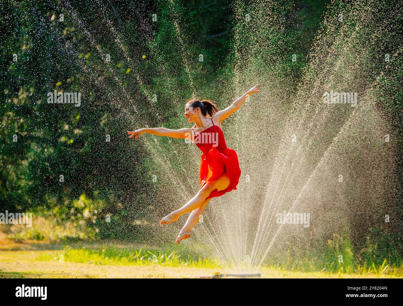 Une ballerine dans une robe rouge effectue un saut gracieux au milieu d'un jet d'eau avec la lumière du soleil filtrant à travers. Banque D'Images