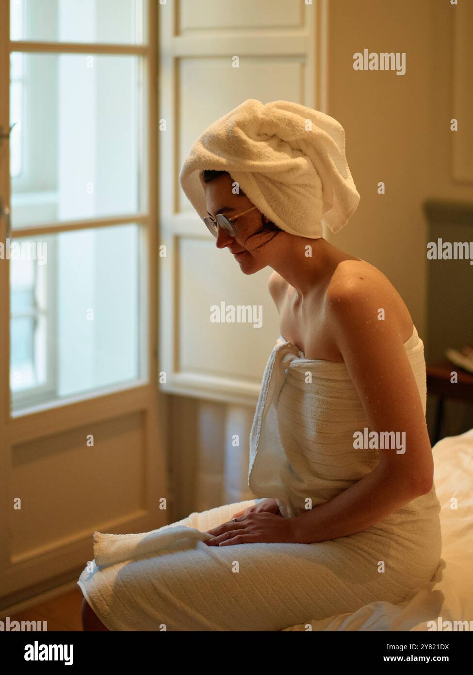 Femme enveloppée dans une serviette assise à côté d'une fenêtre en contemplation après une douche, avec une expression sereine. Banque D'Images