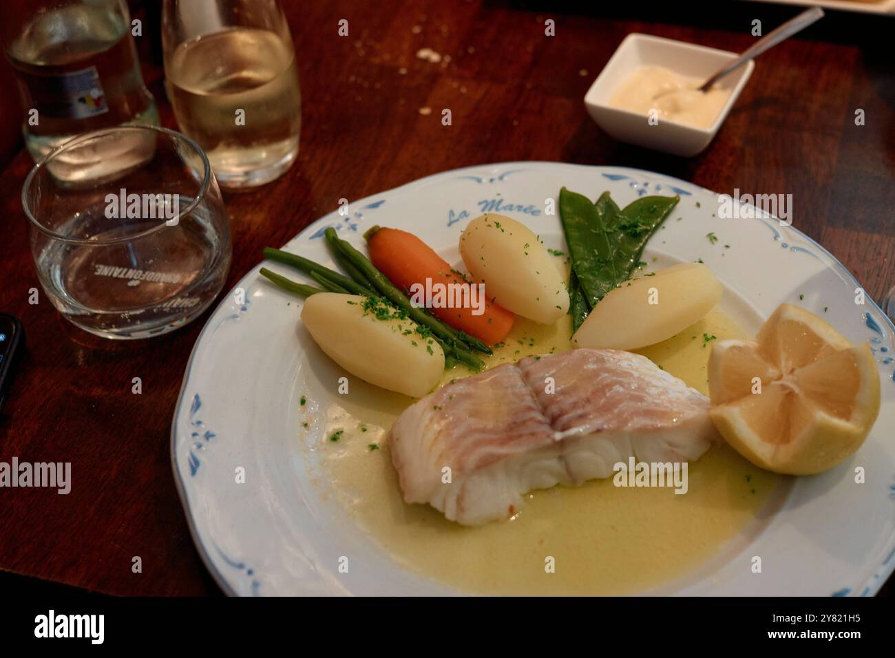 Un repas de poisson poché avec citron, pommes de terre bouillies, carottes et épinards servi sur une assiette blanche avec un verre d'eau et de vin blanc sur une table en bois. Banque D'Images