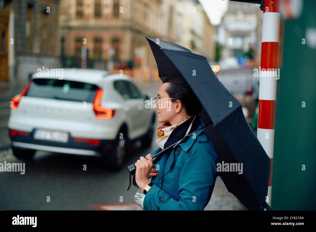 Personne adulte attendant de traverser la rue par un jour nuageux, tenant un parapluie noir ouvert. Banque D'Images