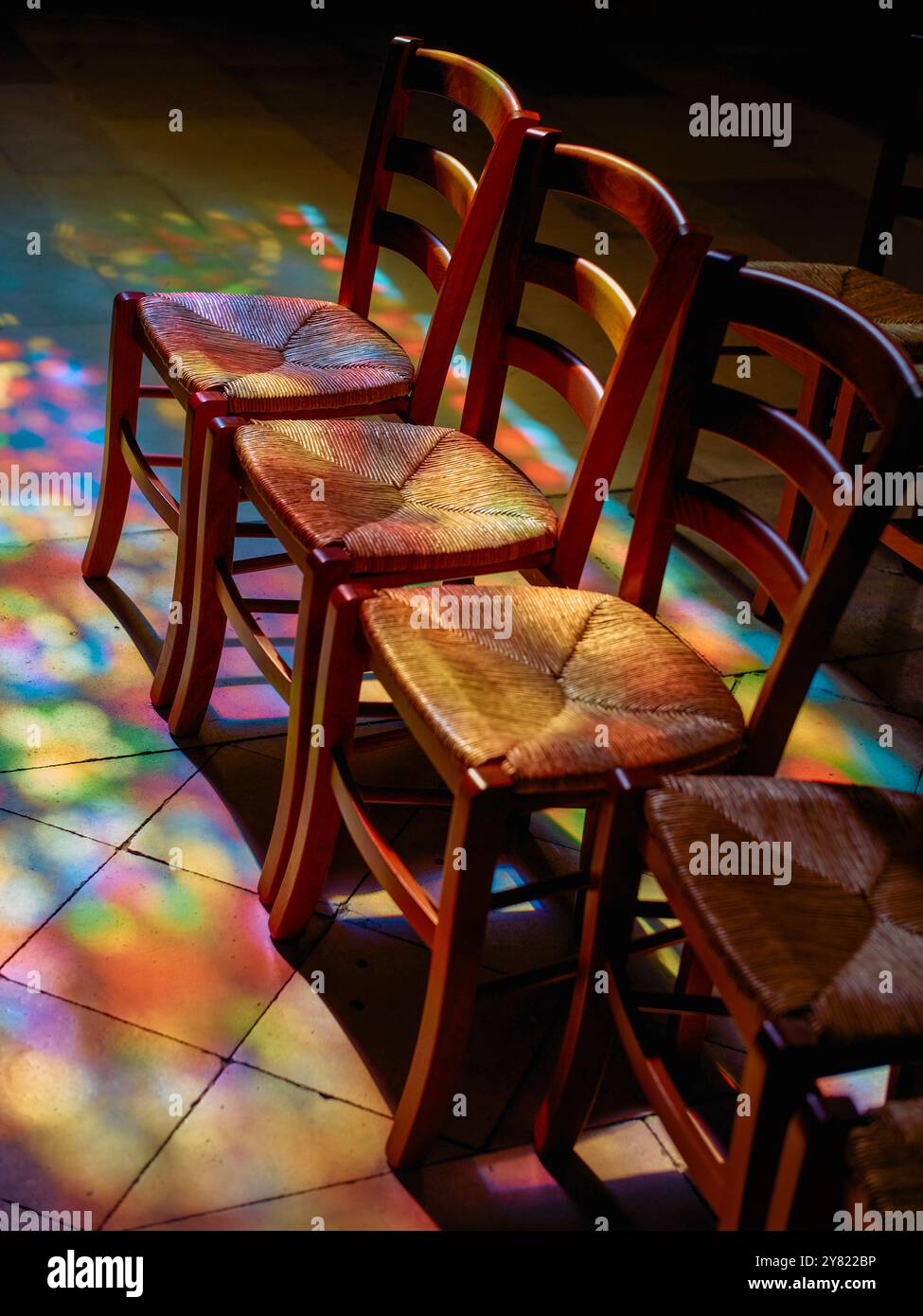 Motifs lumineux colorés sur le sol projetant une lueur chaude sur une pile de chaises en bois. Banque D'Images