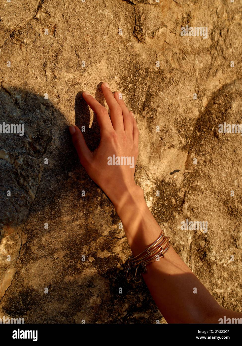 Une main avec des bracelets en argent tendre la main pour toucher une surface de roche texturée pendant la lumière du soleil d'heure dorée. Banque D'Images