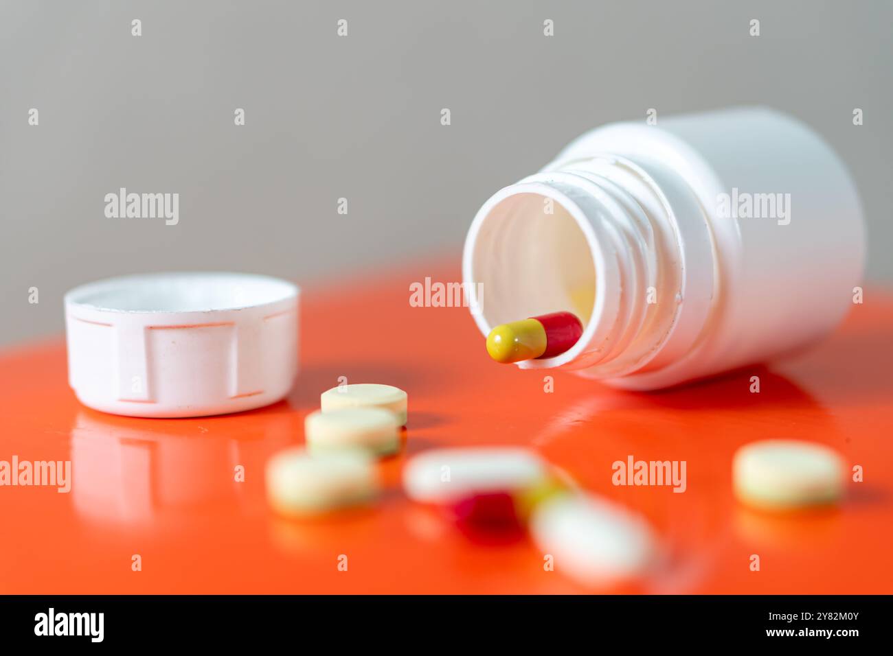 Une bouteille de pilule blanche avec son bouchon enlevé, couché sur le côté avec diverses pilules et capsules se renversant sur une surface orange. Banque D'Images