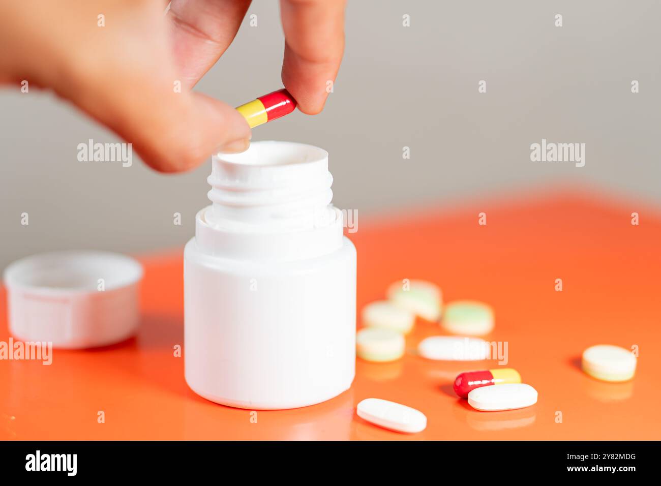 Une main tenant une capsule rouge et jaune sur une bouteille de pilule blanche avec diverses pilules dispersées sur une surface orange. Banque D'Images