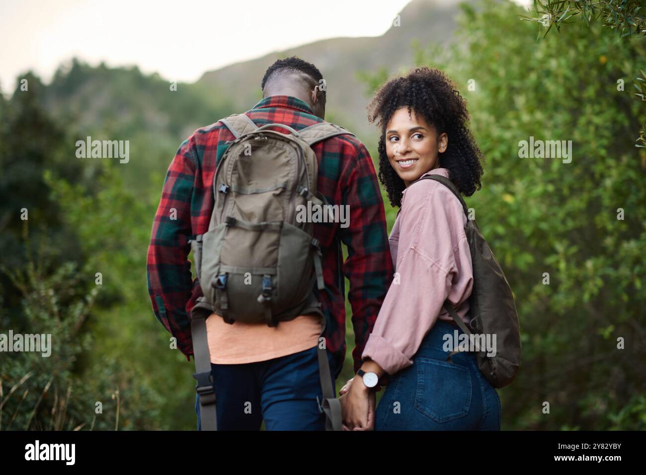 Souriant jeune couple multiethnique tenant la main alors que dehors pour une randonnée ensemble le long d'un chemin dans quelques collines accidentées en été Banque D'Images