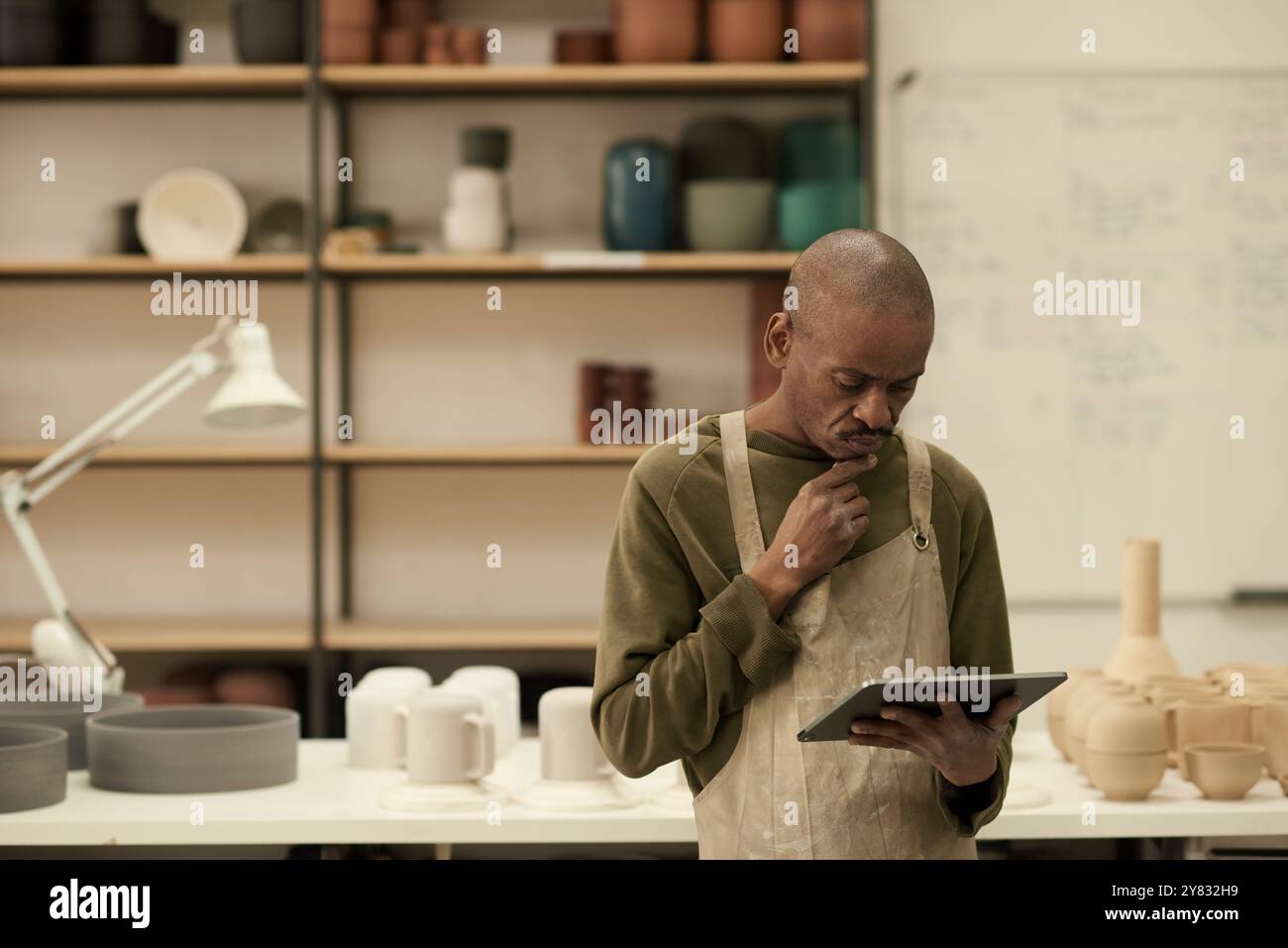Responsable d’atelier de poterie africaine travaillant sur une tablette numérique tout en se tenant debout dans un grand studio de fabrication de céramique Banque D'Images