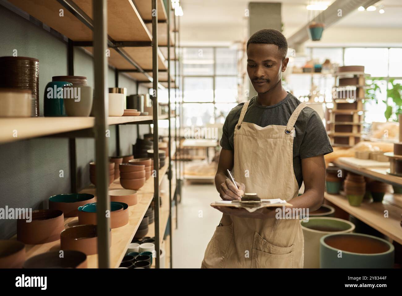 Jeune ouvrier de studio de poterie africaine avec une planche à pince faisant l'inventaire des bols assis sur les étagères dans un atelier Banque D'Images