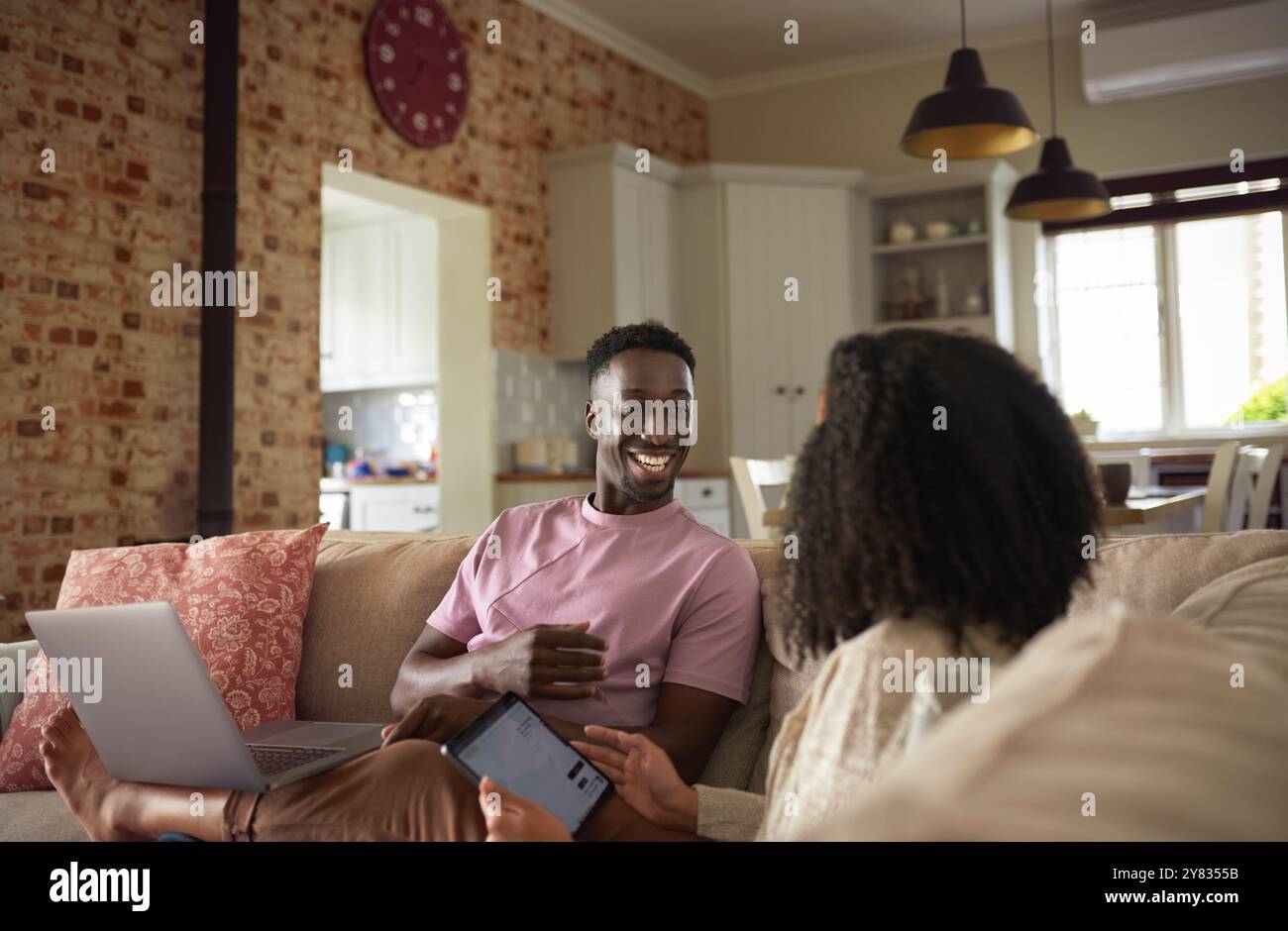 Rire jeune couple multiracial utilisant un ordinateur portable et une tablette numérique tout en se relaxant ensemble sur leur canapé de salon Banque D'Images