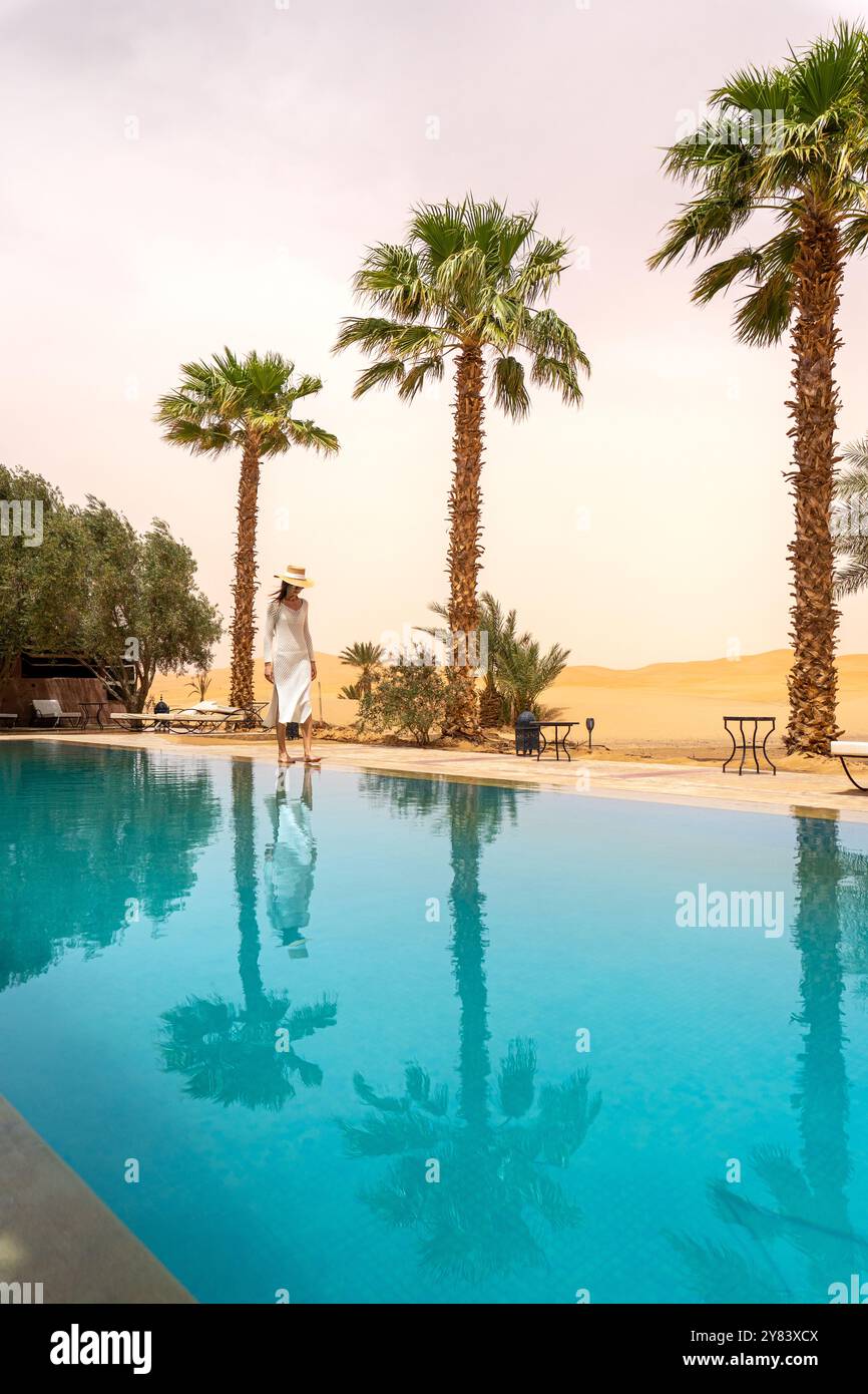 Jeune femme en robe blanche et chapeau debout près de la piscine à la base des dunes du désert de Merzouga Banque D'Images