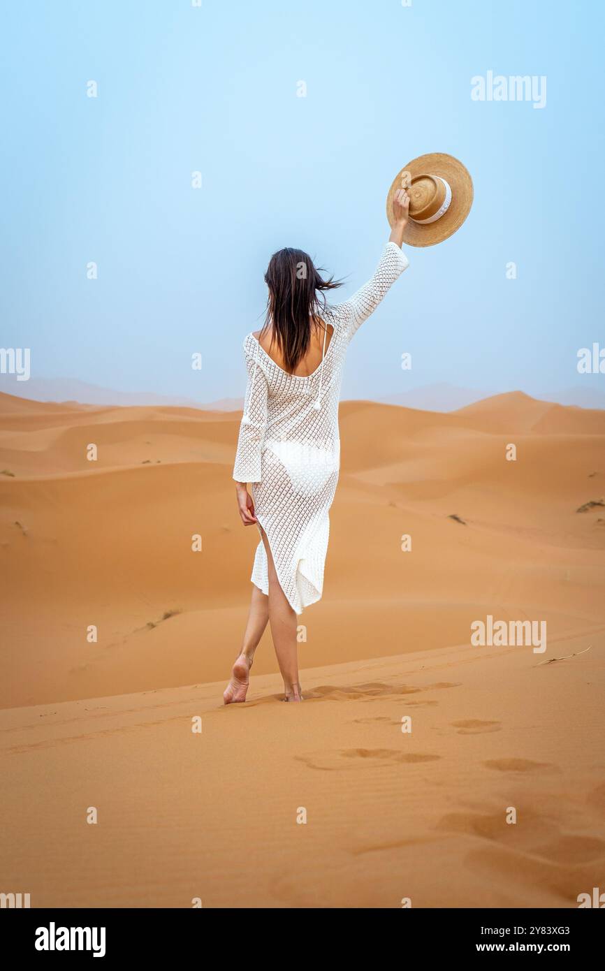 Jeune femme Brunette en chapeau de chameau et robe blanche ondulant de derrière dans les dunes Erg Chebbi Banque D'Images