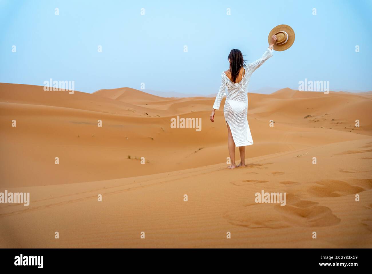 Jeune femme Brunette en chapeau de chameau et robe blanche ondulant de derrière dans les dunes Erg Chebbi Banque D'Images