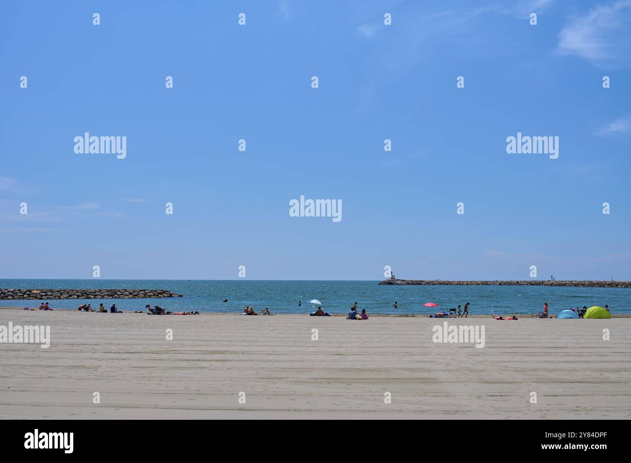 Les gens se détendent sur une plage ensoleillée surplombant la mer Méditerranée calme et un brise-lames sous un ciel bleu clair, Saintes-Maries-de-la-mer, Camargue, F Banque D'Images