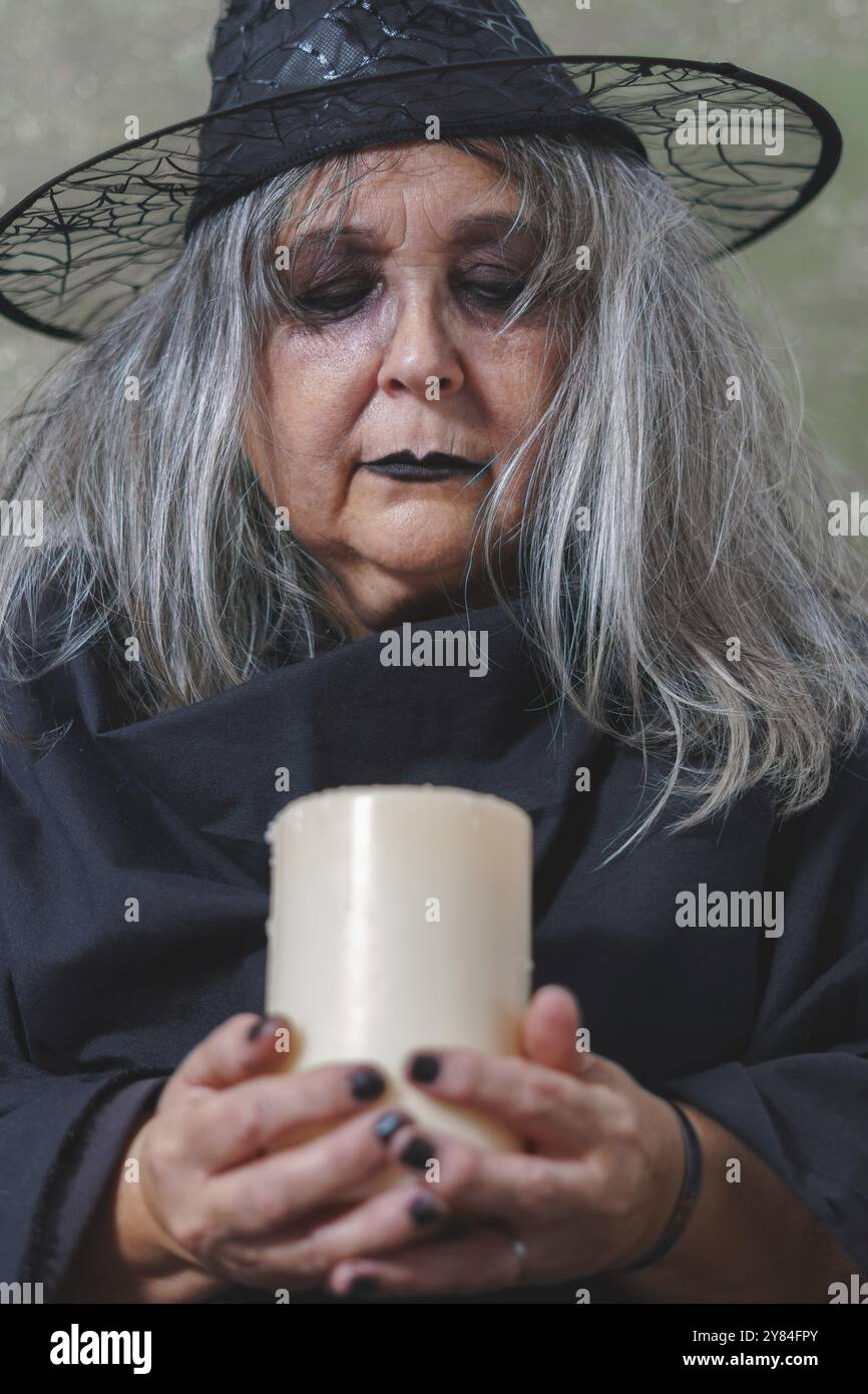 Gros plan d'une vieille sorcière aux cheveux blancs vêtue de noir avec un chapeau et une grande bougie blanche dans ses mains Banque D'Images